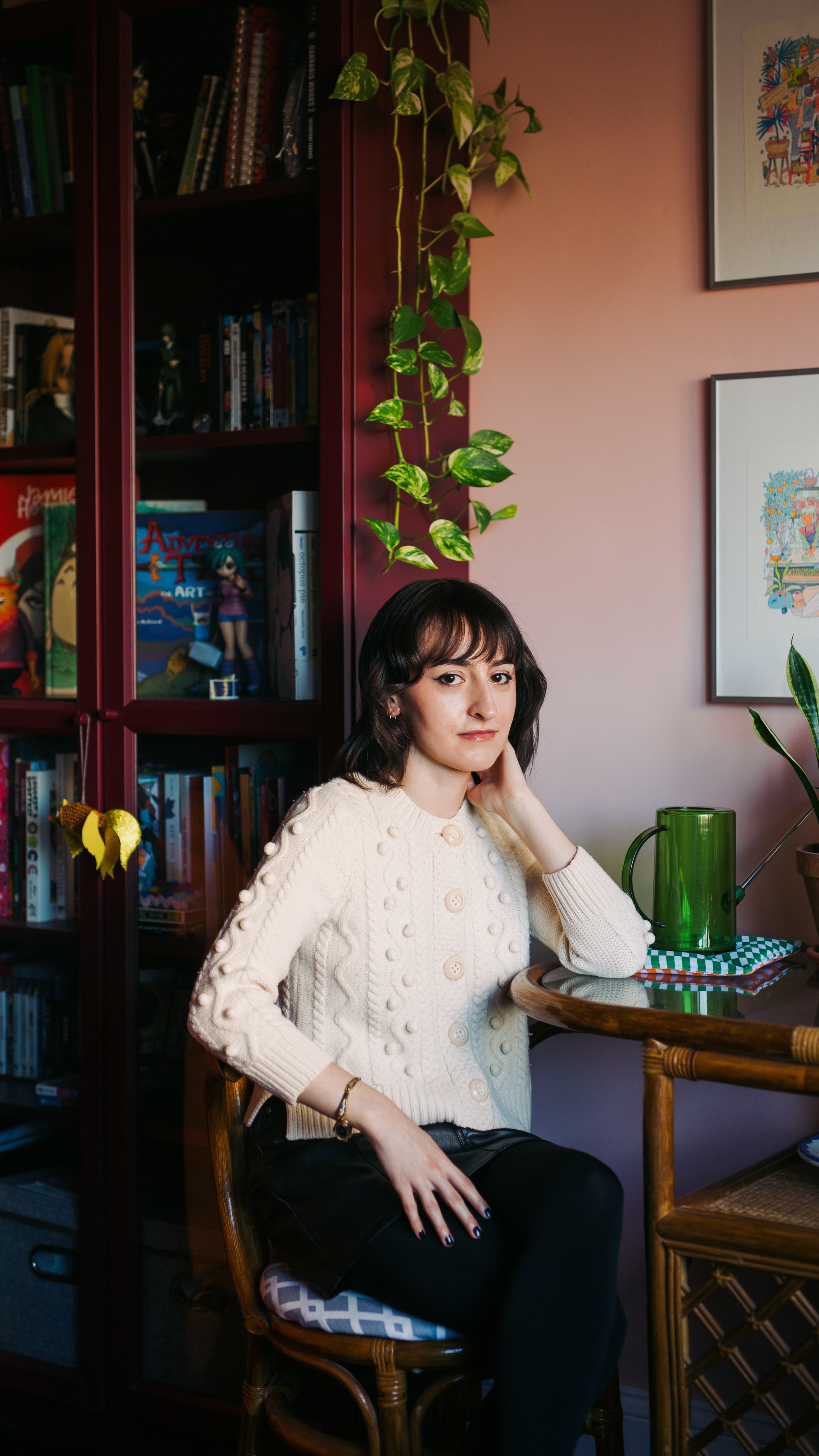 Woman in a cream cardigan sits at a table beside a red bookshelf and plant.