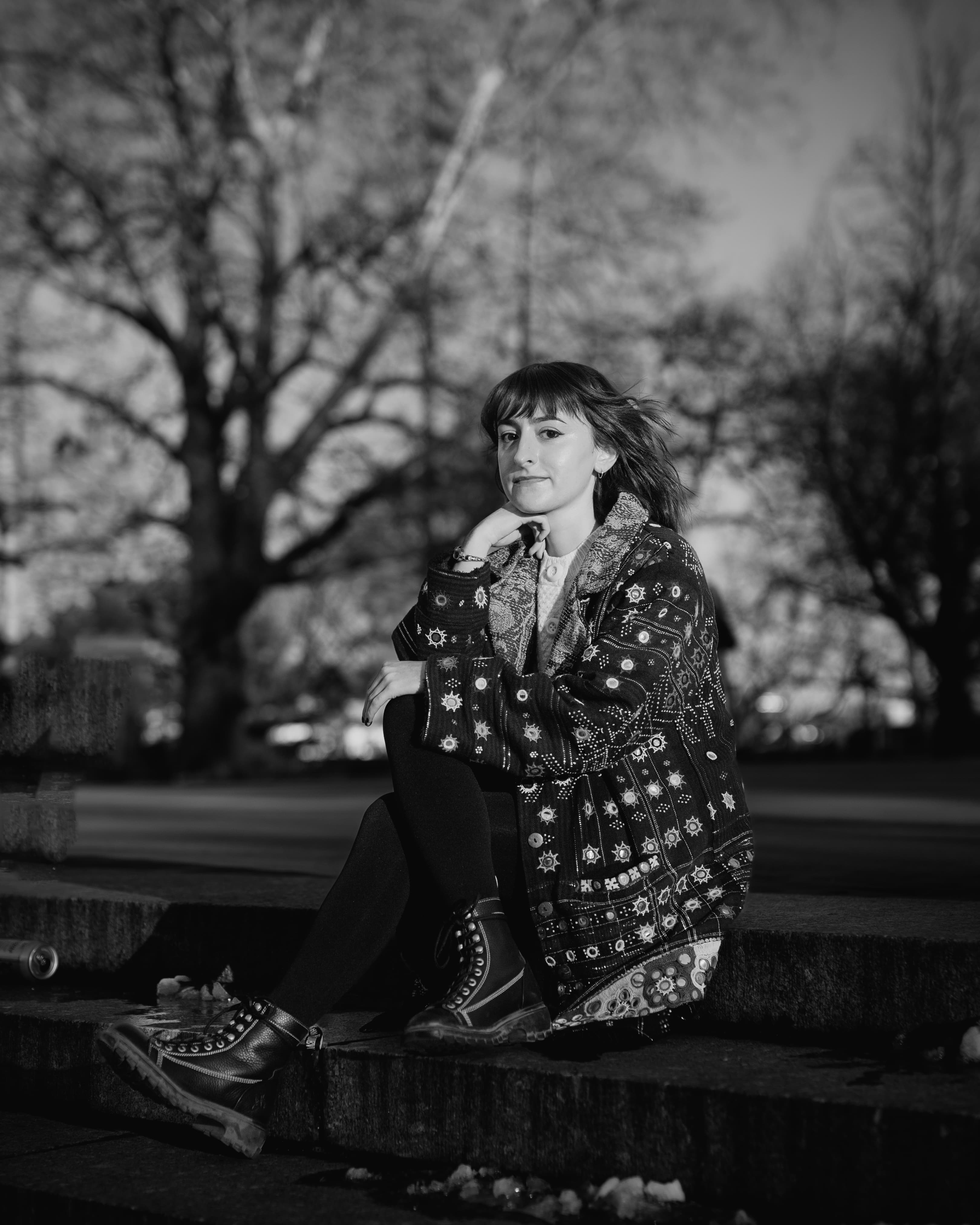 Woman in a patterned coat and boots sitting on stone steps in black and white.