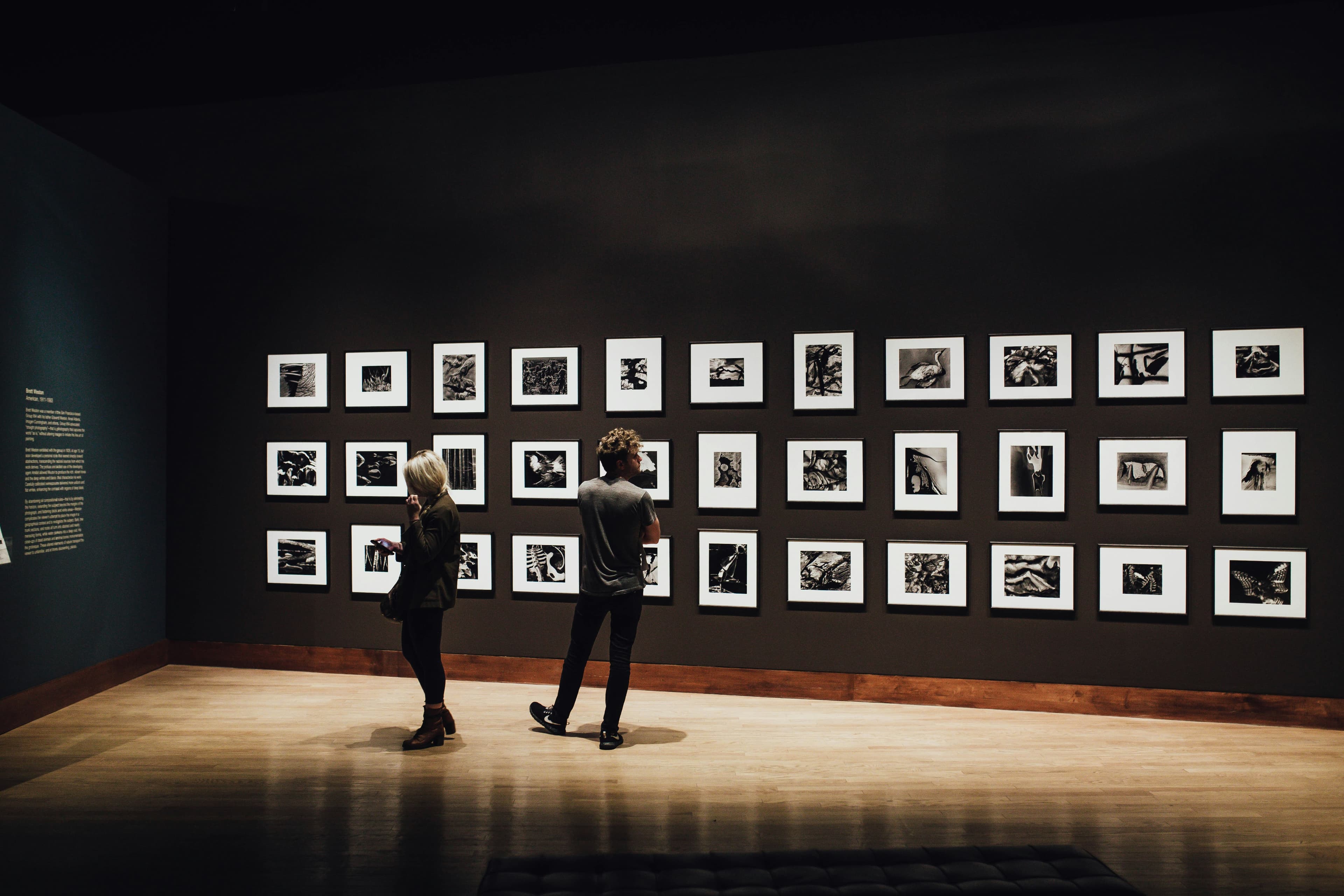 Two people view a grid of framed black and white photographs on a dark gallery wall.