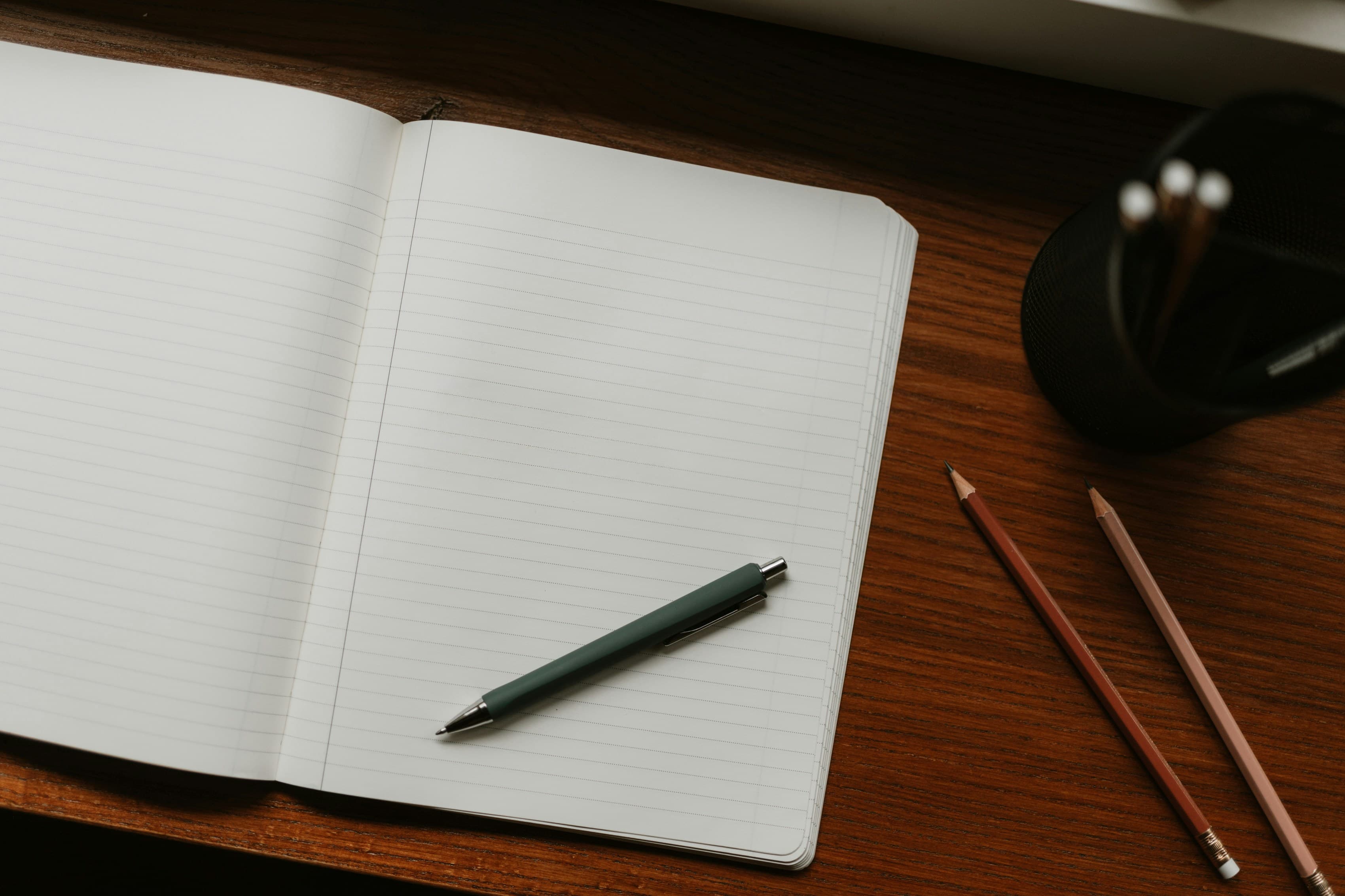 Open blank lined notebook with a green pen and pencils on a dark wooden desk.